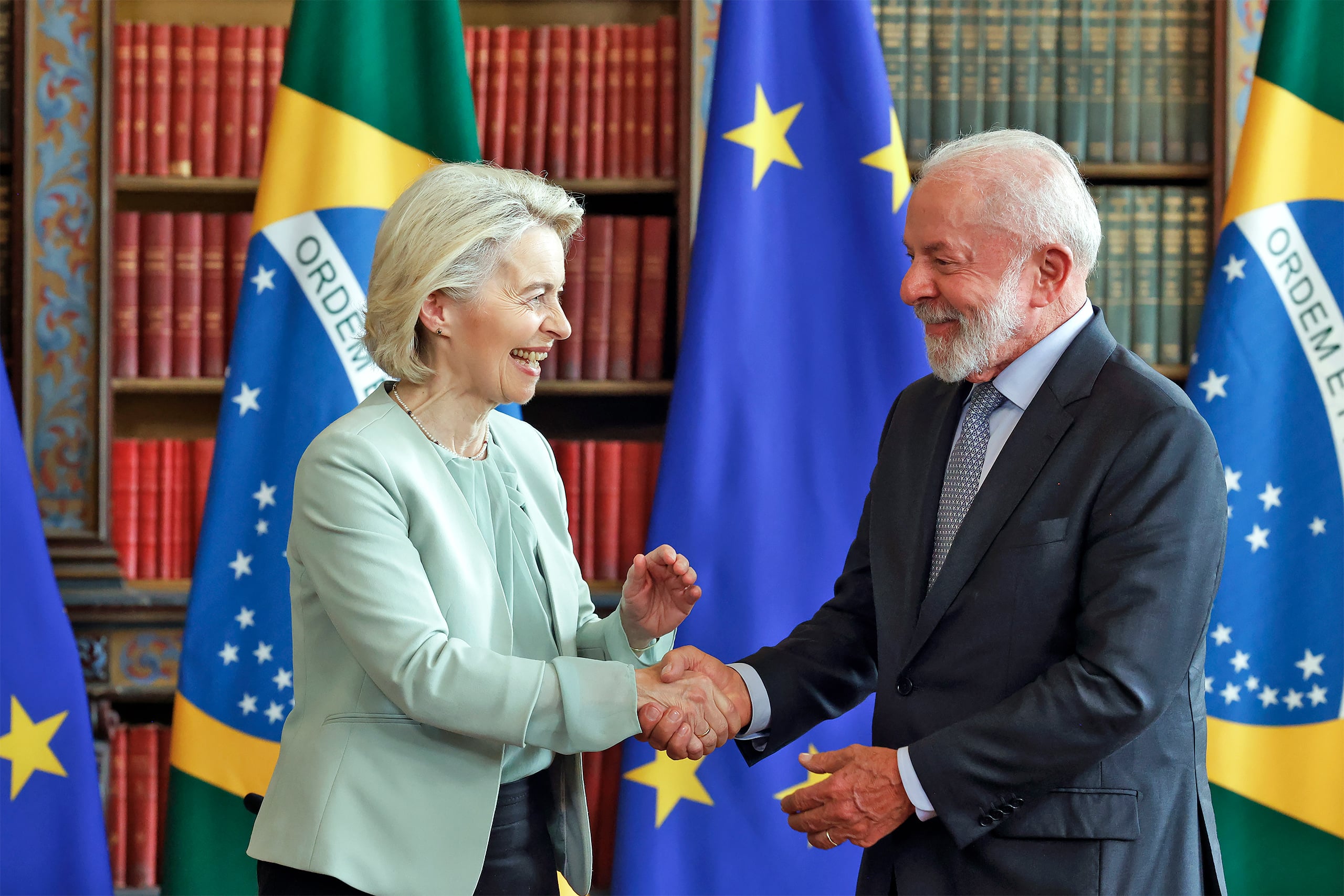 Dos funcionarios se dan la mano durante una ceremonia formal, rodeados por las banderas de Brasil y la Unión Europea